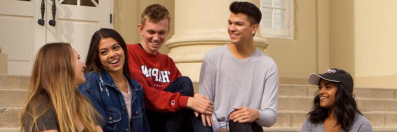 five students sitting in front of a building smiling and talking